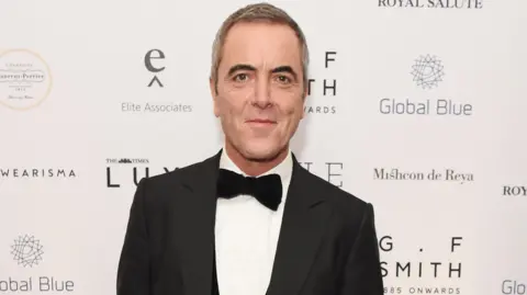 James Nesbitt wearing a black tux with a black bow tie. He's standing behind a backdrop at a red carpet event. 