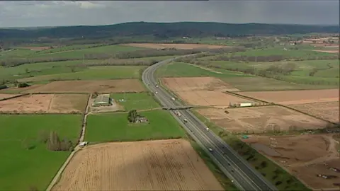 A drone shot of the proposed site of Marlcombe near Exeter Airport. It's a rural area with grass and farm land along with a busy road with two lanes in each direction. 