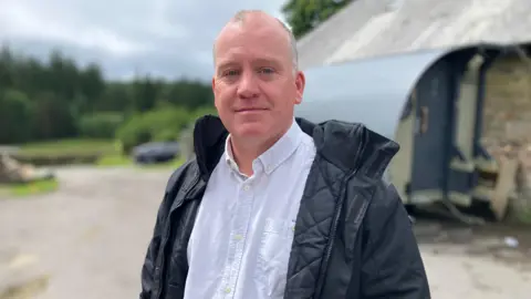 Phil Maddock in a white shirt and black jacket standing outdoors near a building, with a rural landscape in the background.
