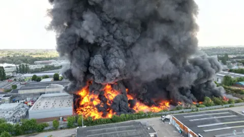 Steve Hubbard/BBC A huge plume of black smoke and flames pour from the warehouse site, with other industrial buildings visible in the background.