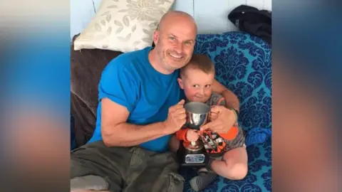 Kevin cuddles his son Benny on a blue sofa. They both hold a trophy cup. 