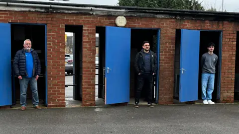 Emma Baugh/BBC Michael, Ollie and Oscar, are stood in a line. Each one is standing in a different turnstile doorway. The building is outside and red bricked with blue doors, which have been opened. The spaces in which they are stood is where they go to check tickets as fans come into the stadium. 