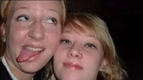 Becca Hainsworth A close-up image of two young women looking into the camera. The woman on the left is looking off to the side and sticking her tongue out. The woman on the right is looking into the camera. 