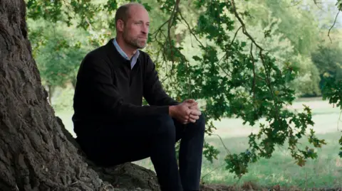 PA Media Prince William sits at the foot of a tree looking out beyond the shot of the camera.