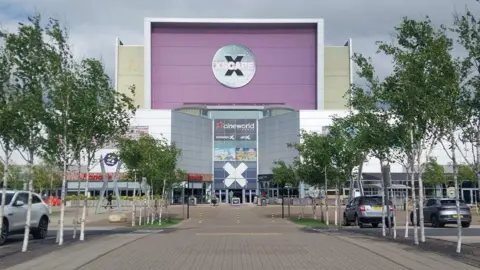 The front of the Xscape leisure complex in Castleford, pictured from the car park. The entrance has several tiers. A series of planters and trees are outside and a number of cars are parked.