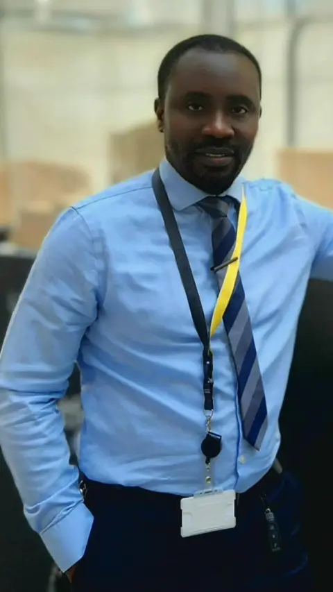 Titi Owode Titi looking at the camera, wearing a smart blue shirt, a striped tie and trousers.