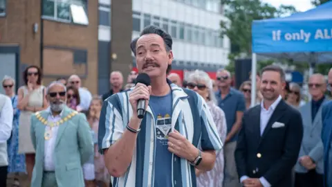 Dartford Borough Council Craig Revel Horwood holding a microphone. There is a crowd of people behind him