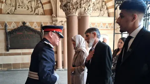 Jenny Kirk/BBC A police official wearing a hat and a police uniform greets members of a family at the entrance of a large ornate civic building. The officer speaks to a man in front of him who rests one hand on his chest. Other members of the family stand alongside him.