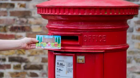 A hand holding a postcard with an illustration of a palace on it reaches towards a red postbox.