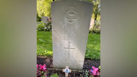 Chris Bullock A close-up of a Commonwealth War Graves Commission headstone in pale stone. It has the RAF wings at the top and is inscribed with Wilfred Surplice's name and a cross. Behind it is grass and more gravestones. In front are flowers and a small wooden cross placed in the ground.