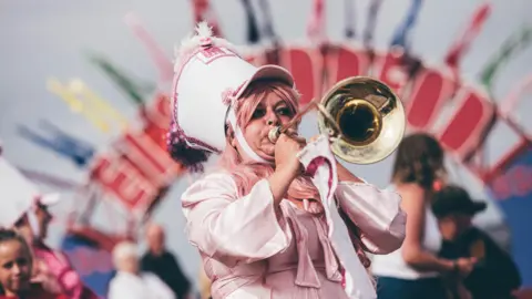 Aled Llywelyn | Eisteddfod Genedlaethol A woman wearing a pink costume and a pink parade hat blows into a trumpet.