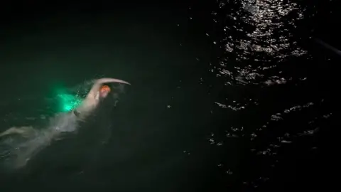 Grimsby & Cleethorpes Water Rats A man is mid-action swimming freestyle through the dark sea with a green light attached to him. He is wearing an orange swim cap.