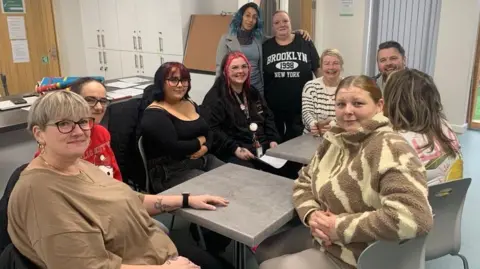 MICKLUNNEY/BBC A group of people sit around two small tables in a cafe. Two people stand at the back. They smile at the camera.