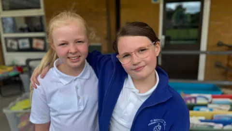BBC Mali and Ffion stand arm in arm in a school classroom. The two girls are wearing white polo shirts, one with a blue fleece with the school logo on. One of the girls has long blonde hair tied back in a ponytail, the other has brown hair tied back and wears glasses.