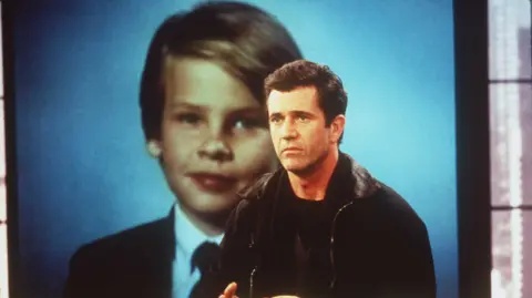Touchstone Pictures/Getty Mel Gibson acts as the character in Ransom, a father whose son has been taken hostage. He looks to one side of the shot, with a large screen behind him showing a photo of his son, who is aged about 8 and wears a suit. Mel Gibson wears a black t-shirt, sweater, and black jacket.