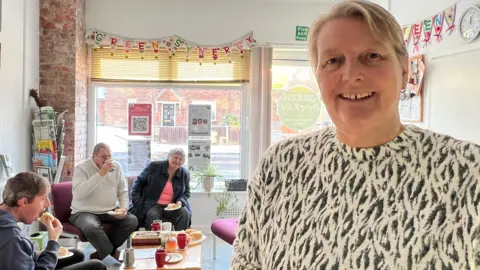 Wendy Slater is wearing a black and white patterned top and is smiling. She has blonde hair. Behind her are three people sat on chairs eating sandwiches. Colourful bunting lines the room. 