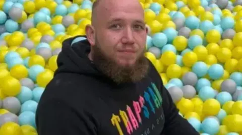 Supplied Picture of Chad Allford sat in front of a ball pool wearing a black hoodie. He has a brown beard and a shaven head.