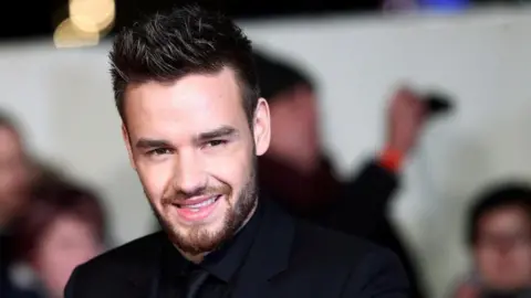 Reuters Liam Payne, dressed in a black shirt, suit and tie, smiles for photographers at a red carpet event with several onlookers behind him