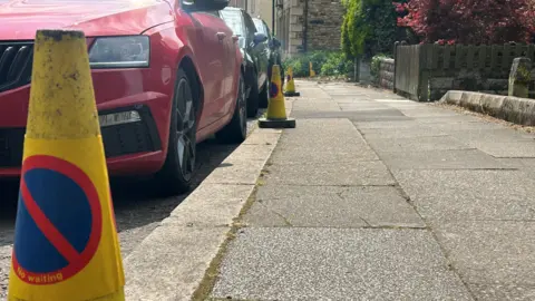 Traffic cones lining a pavement with three cars parked along the kerb.