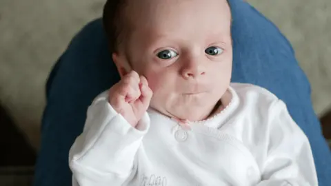 Elizabeth Bull A small baby lying on a person's knees. The baby is in a white baby grow with one hand clenched and the baby's lips are tightly closed. 