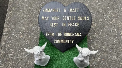 A heart shaped sign saying: 'Emmanuel & Matt May Your Gentle Souls Rest In Peace From The Buncrana Community' with two angel statues next to it.