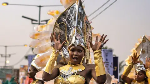 Olympia De Maismont / AFP A musician wearing a gold dress with gold glove sleeves and a large silver headpiece performs jazz hands during the Calabar Carnival