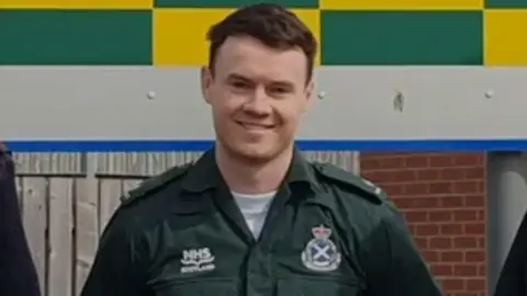 Stephen Doohan smiles at the camera. He has short brown hair and wears a green NHS paramedic uniform
