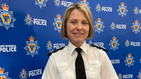 Northumbria Police Chief Constable Vanessa Jardine standing in front of a banner displaying the force's insignia. She is wearing the white shirt and black tie of her uniform and has blonde hair cut into a bob.