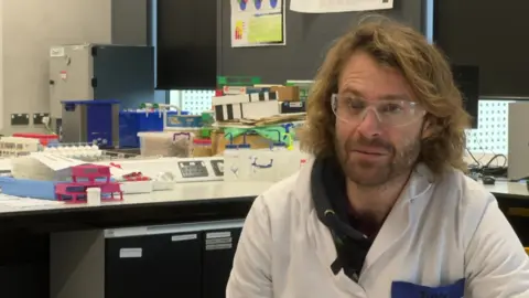 Dr David Megson, who is wearing a white lab coat and protective glasses, is sitting in laboratory. 