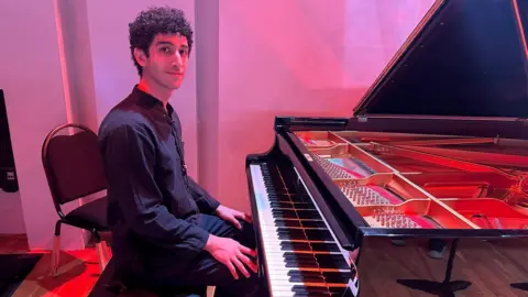 Cad Taylor/BBC A young man sits at a piano with performance lighting in the background