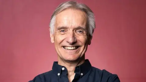 Phil Powell wearing a blue shirt smiling at the camera, against a red backdrop 