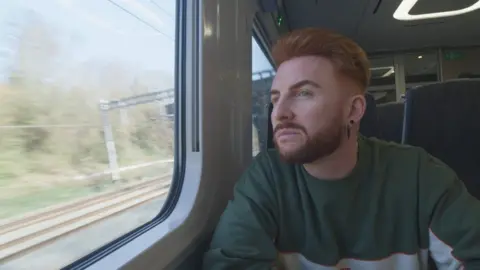 Wegrzyn has a ginger quiff and beard and looks out of a train window as it moves down the tracks, deep in thought. They have two black earrings in their ears and are wearing a green and grey jumper. 