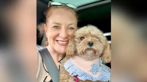 Family photograph Selfie of Jacqui Padden with long dark red auburn hair tied up in a ponytail wearing sunglasses propped on her head and a beige and black cardigan with dog, Wilma, in a car. She is smiling as she holds her minature poodle.