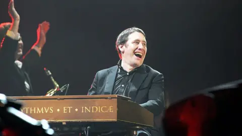 Getty Images Jools Holland wearing a black suit laughing on stage sat at his piano