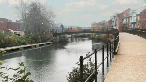 Stantec Image shows a footbridge with a cyclist going across it, linking a housing estate to a shopping centre. In the image you can see a concrete pathway and river running beneath the bridge.