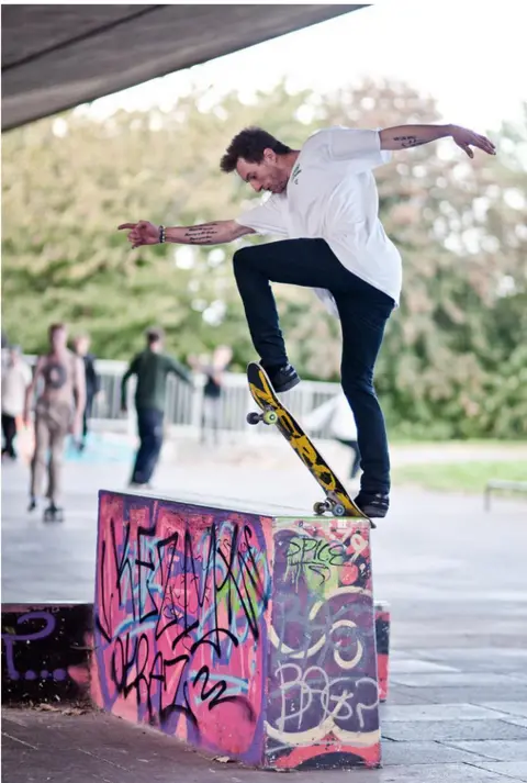 Johnny Haynes A male skateboarder doing a move on a small block that is painted in graffiti, largely pink and purple. It is under the flyover. Others are skateboarding in the background. Trees are out of focus in the background too.
