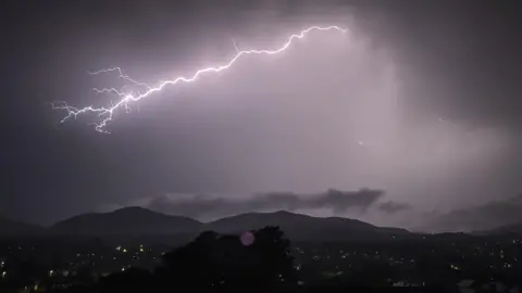 BBC WEATHER WATCHERS - ALICE EA. Cielo nocturno oscuro con un brillante relámpago. Al fondo se ven colinas oscuras.