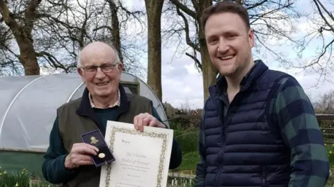 David Maxwell Two men are standing in an allotment. The man on the left is holding a certificate in one hand and a medal in the other hand. To his right is a man wearing a blue body warmer and chequered shirt. They are both smiling at the camera.