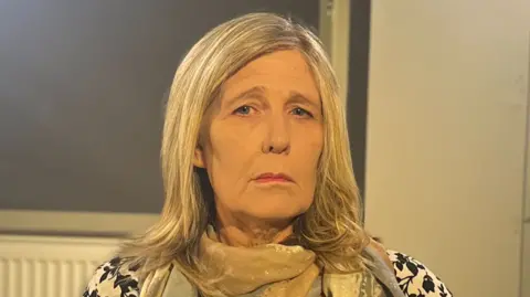 Vanessa sits in front of a white wall. She is an older woman with straight, shoulder-length blonde hair and is wearing a white top with black flower pattern.