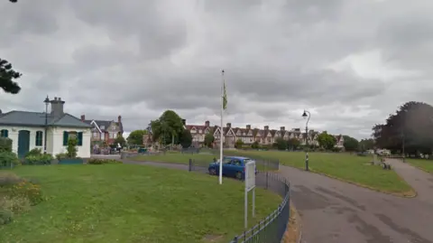 A google street view image of Freedom Fields Park in Plymouth. There is a white building in the left. There are big areas of green grass with concrete footpaths. There is a blue car parked. There is a blue fence around the grass outside of the white house. There are green lamp posts. There are brown houses in the background. 