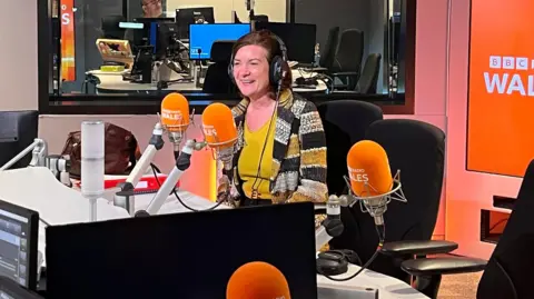 Eluned Morgan sat wearing headphones in a BBC Radio Wales studio, in front of a microphone branded with the station's logo.