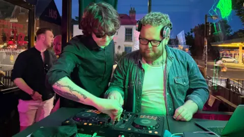 Two young men stand over a DJ turntable, turning dials, both focused on the decks. The man on the left wears a black-buttoned shirt, black sunglasses and has long dark hair. The man on the right wears a blue denim shirt, with a white t-shirt underneath. He has a ginger beard and black-framed glasses. Behind them is a pub setting, and out of the window a garage, cars on the road and other buildings can be seen.