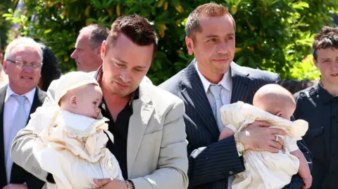 PA Media Barrie Drewitt-Barlow is wearing a cream suit jacket over a black shirt, while Tony has a pinstriped navy suit on over a white shirt and pale blue tie. The babies are wearing frilled white outfits and are being carried by their parents. It is a happy scene on a sunny day outdoors.