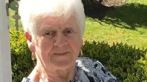 Family photograph Marie Cunningham with white hair and gold hoop earrings wearing a black-and-white patterned blouse. She is sitting in a garden.