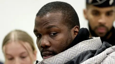 Reuters Mathias Pogba, brother of Paul Pogba, wearing a winter coat, looks to the side of the camera as he arrives to attend the verdict in his trial. Two other people are out of focus in the background behind his head. 