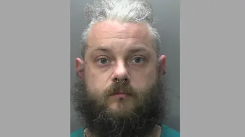 A mugshot of Jamie Elliott following his arrest. Jamie has greying hair, a big beard and is wearing a nose ring. 