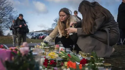 Two women crouch beside a memorial for the victims of a mass shooting in Sweden. It's daylight, and you can see the women lighting candles and placing them beside bouquets of flowers.