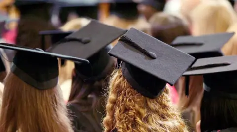 Students sitting with black graduation hats on their heads. The picture is taken from behind them. They have long hair which is blonde, brown or black. 