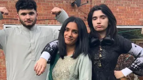 Ayshea Malik, who has shoulder length black hair, stands in a garden in a traditional sari style green dress with her daughter's arm around her shoulders and her son standing on her right with his arms in the air