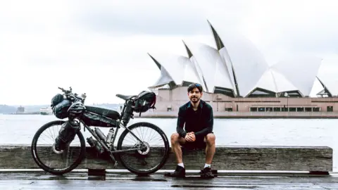 Nathan Hurley A man with dark hair and dark top sits next his bicycle in front of the Sydney Opera House